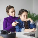 Serious teenage girl doing homework, making research, looking for information online on internet using laptop pc while mother helping her, sitting behind and helping her, pointing finger at screen